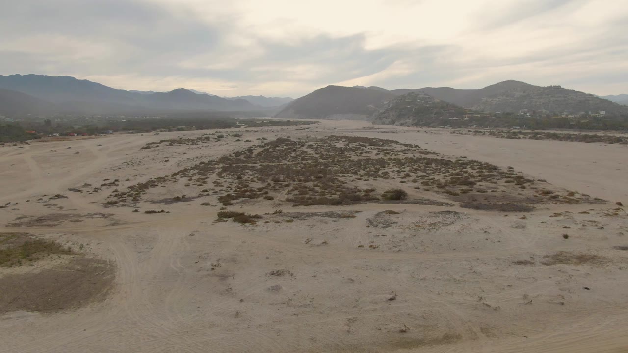 Aerial frame in Los Barriles, Baja California Sur, centered on a sandy rise with patchy low vegetation, open flats around it, layered mountains beyond, cloudy light, and no visible people.