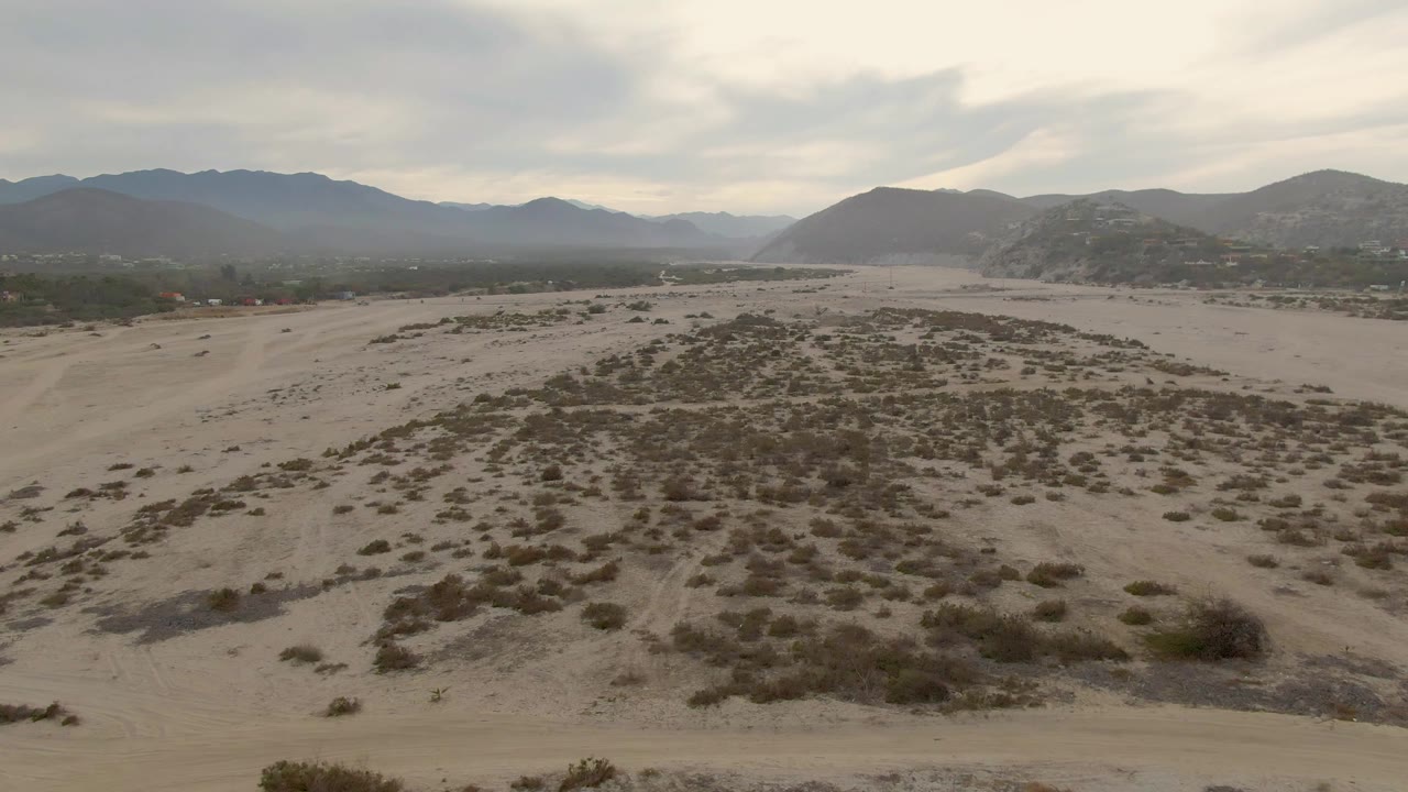 Aerial view over sandy desert scrub near Los Barriles, Baja California Sur, with distant mountain ridges, overcast sky, and no visible people.