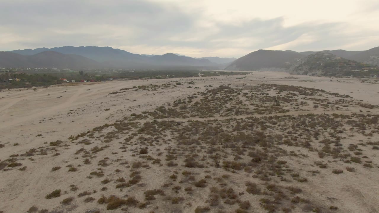Aerial still from Baja California Sur showing scrub-covered sandy ground, a low hill to the right, distant blue-gray mountains, cloudy light, and no visible people.
