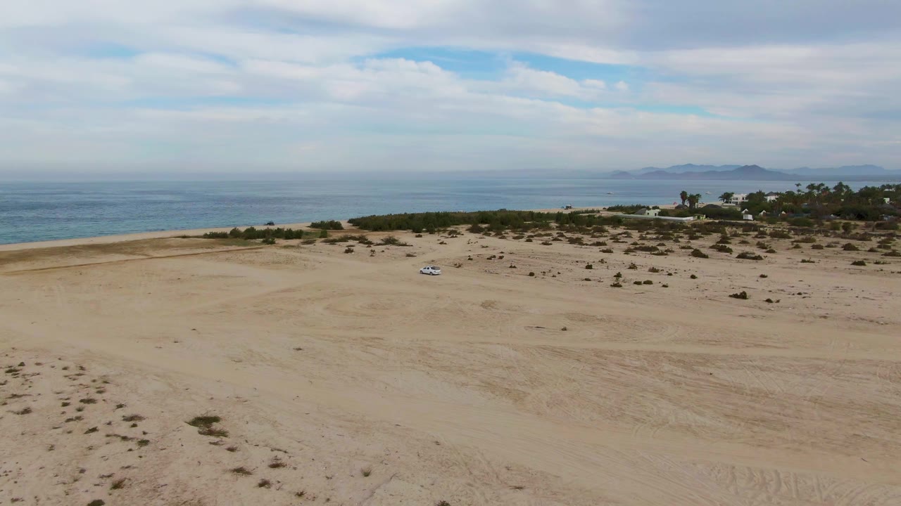 Wide aerial frame over Los Barriles sand flats with a small white car near sparse shrubs, blue ocean on the horizon, distant landforms, and no visible people.