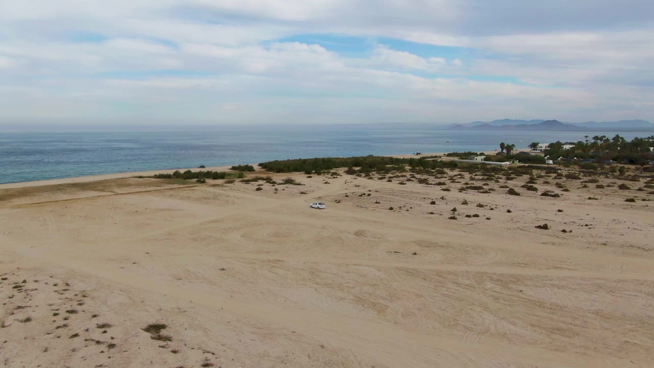 Aerial view in Los Barriles showing the white car farther across the open beach, with dune scrub, a blue ocean band, cloud cover, and no visible people.