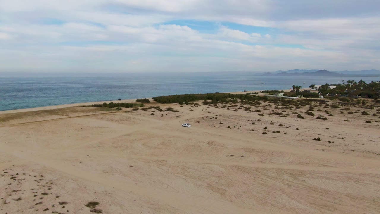Los Barriles aerial frame with the small car near the center of the sandy foreground, low shrubs along the shore, calm blue water beyond, and no visible people.
