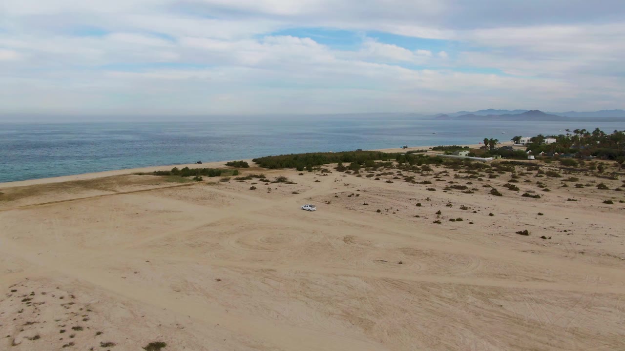 Aerial view over the Los Barriles beach with the car slightly lower in the sandy expanse, scattered scrub patches, blue ocean, distant land, and no visible people.