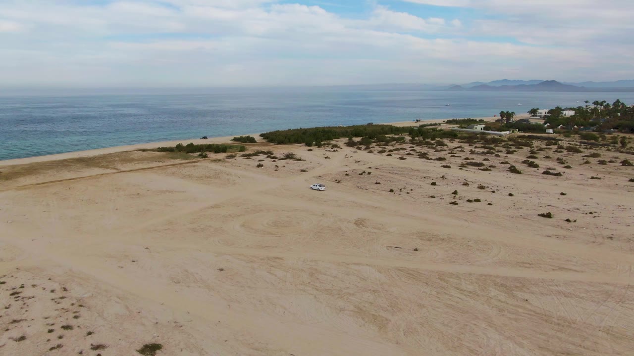 A white car sits small against sandy coastal dunes near Los Barriles, with tire tracks, sparse shrubs, blue ocean, distant land, and no visible people.