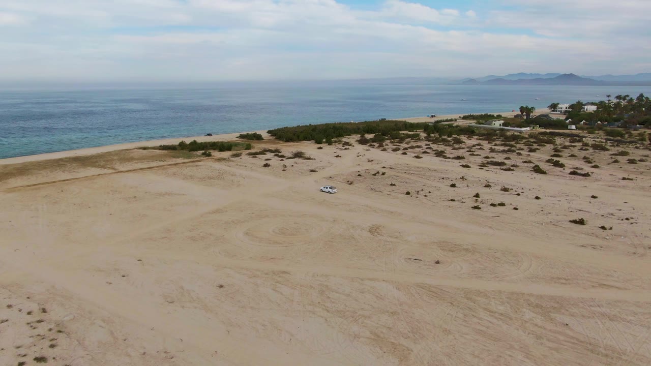 The white vehicle appears farther across the pale sand near Los Barriles, with curving tire marks, patchy shrubs, a beach strip, blue water, and no visible people.