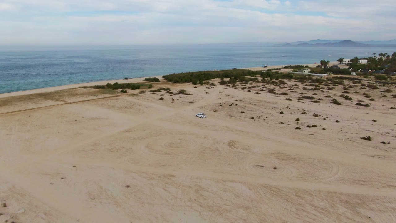An aerial frame shows the white car amid sandy tracks and scattered desert shrubs near Los Barriles, with the ocean and low distant land beyond, and no visible people.