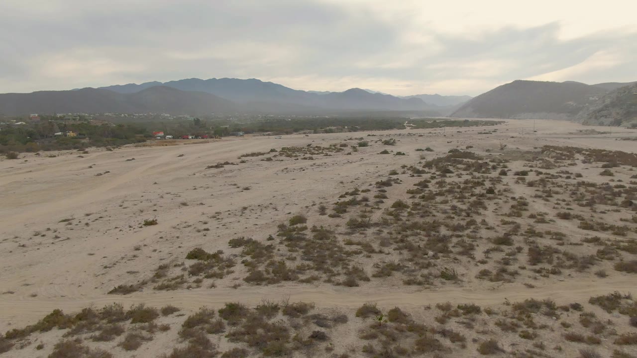 Wide aerial frame over Los Barriles desert plain with scattered shrubs, a faint settlement band, distant mountains, overcast sky, and no visible people.