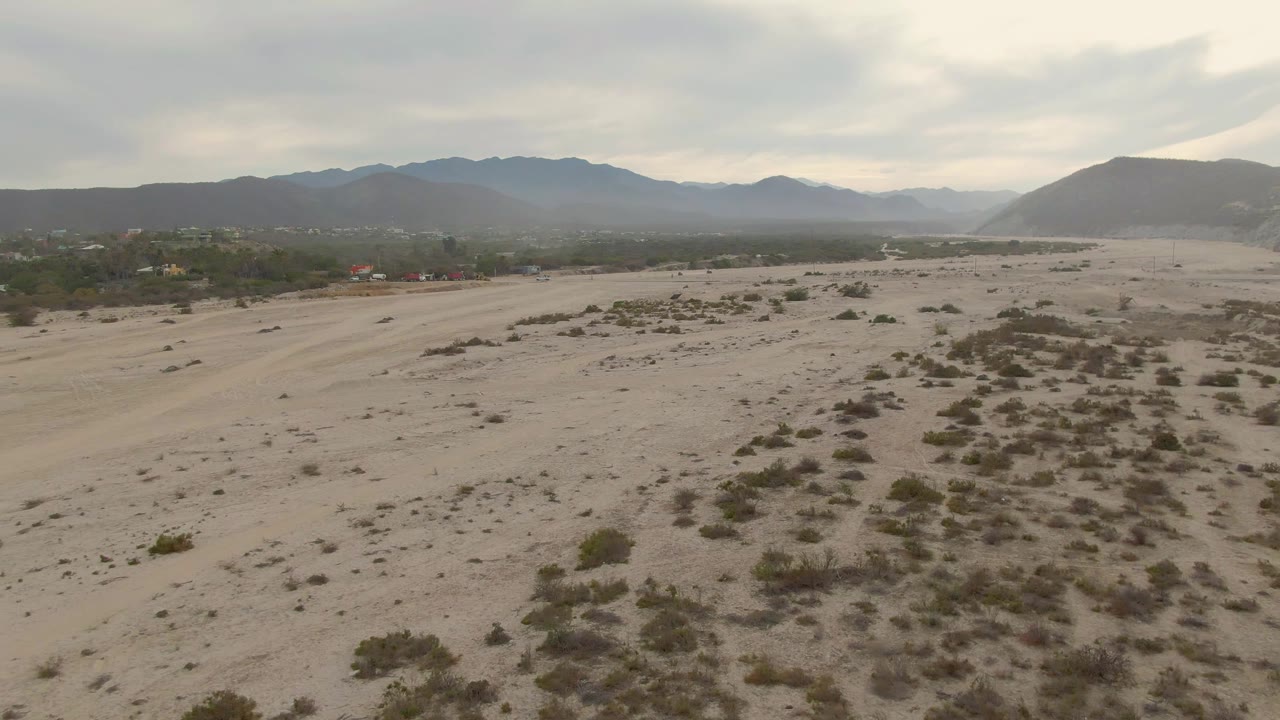 Los Barriles desert aerial with clustered shrubs across pale sand, a low green horizon strip, layered mountains, gray overcast sky, and no visible people.