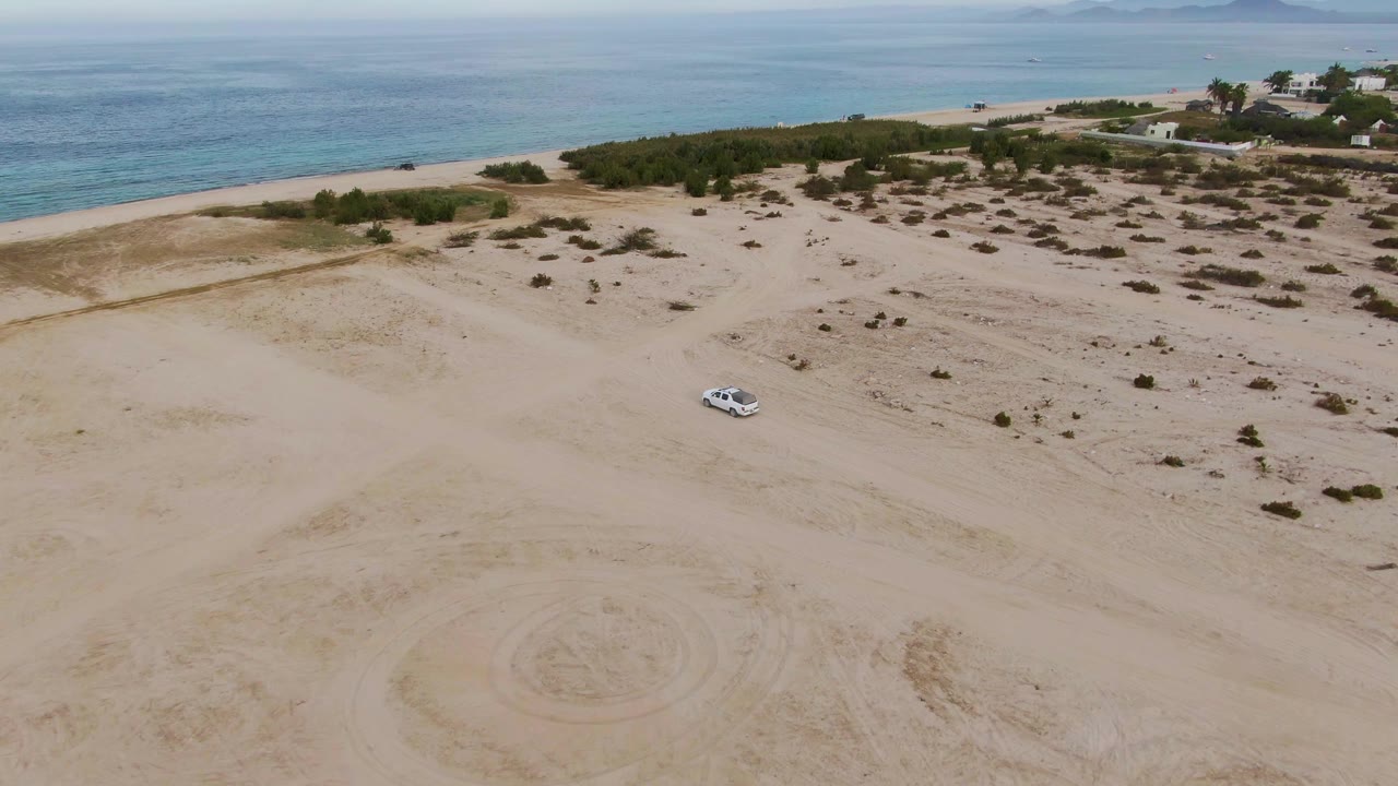 Aerial view in Los Barriles, Baja California Sur of a white car on pale dunes with turquoise ocean, beach vegetation, distant buildings, and no visible people.