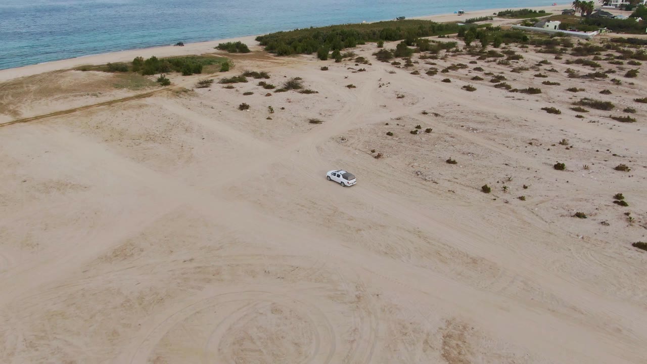 Aerial frame over Los Barriles showing the white car farther across the sandy terrain, with shrubs, tire marks, beach, ocean, and no visible people.