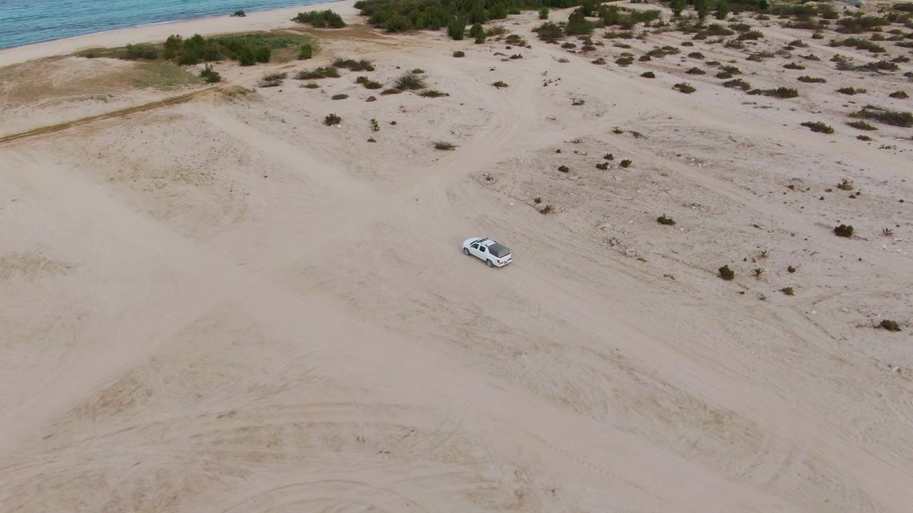 High drone view in Los Barriles with the white car on sandy dunes, small shrubs across the terrain, a strip of beach and ocean at the top, and no visible people.