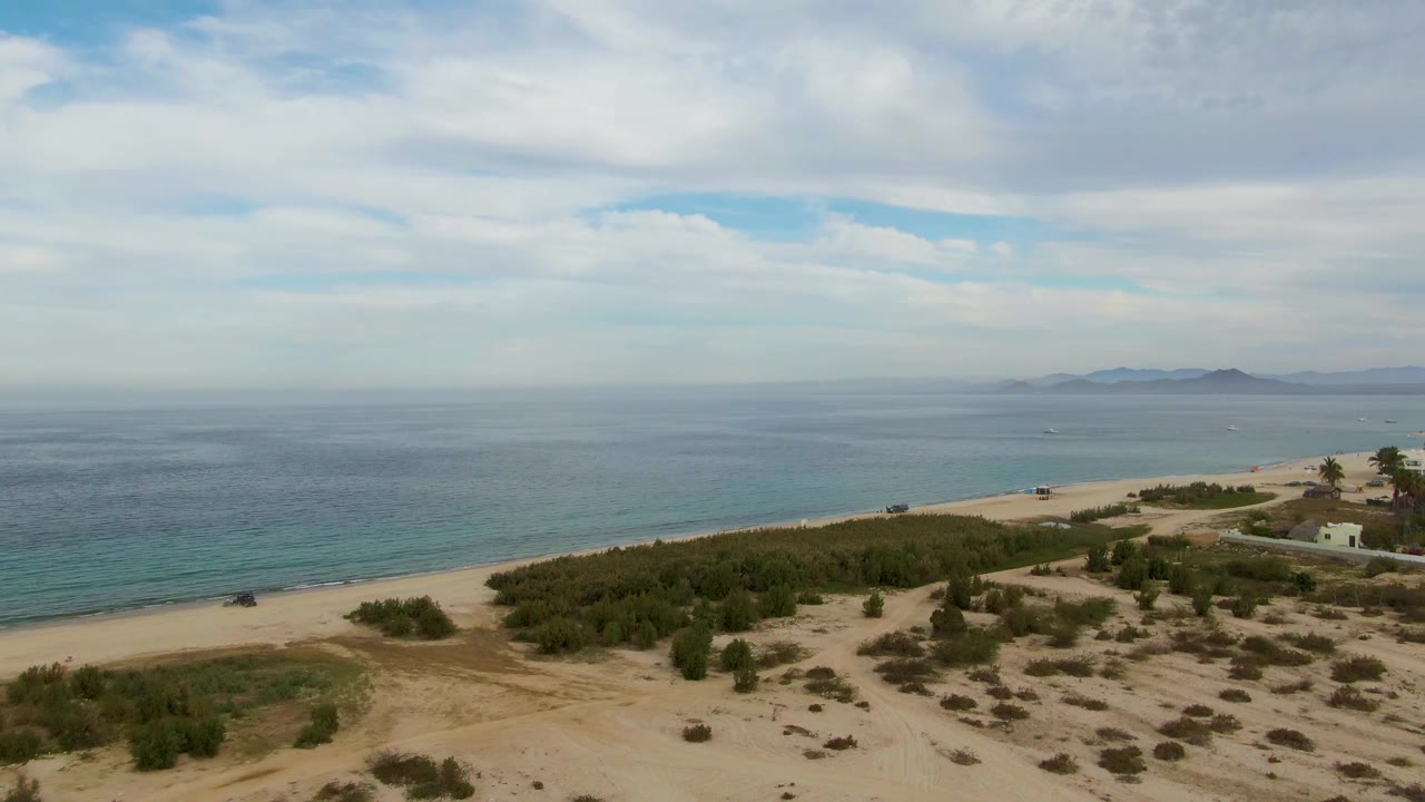 Aerial frame over Los Barriles, Baja California Sur showing sandy dunes, low green vegetation, a shore road, beach, blue ocean, distant land on the horizon, and no visible people.
