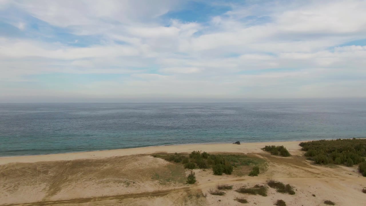 Aerial view in Los Barriles with a broad sandy dune ridge, scattered shrubs, a pale beach strip, and calm ocean water, with no visible people.