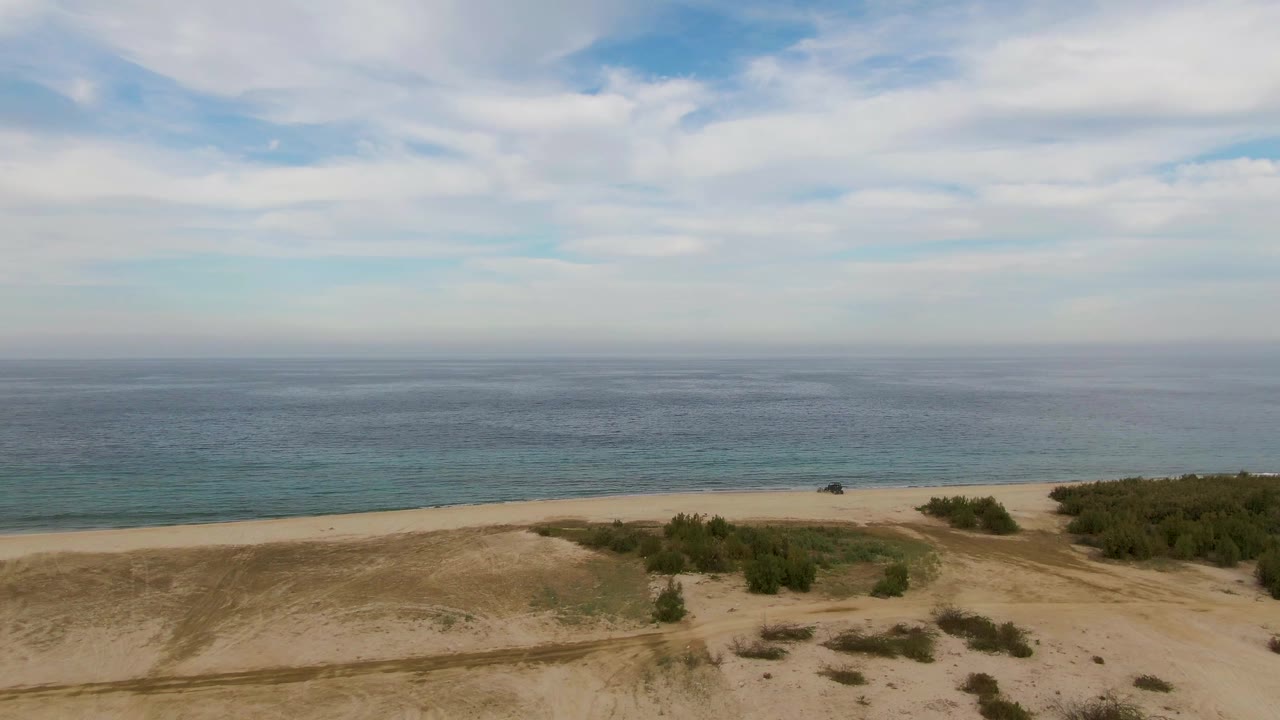 Wide aerial frame of the Los Barriles shoreline with dunes and vegetation patches in front of a long beach and blue ocean, with no visible people.