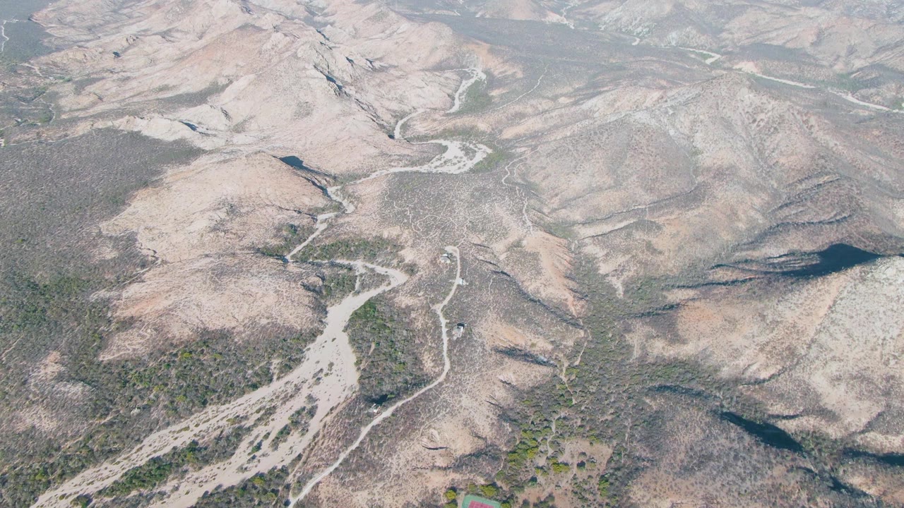 Mid-clip aerial frame over Cabo Pulmo shows a winding dirt track threading between pale rocky hills, green scrub patches, and a small vehicle near the lower roadside, with no visible people.
