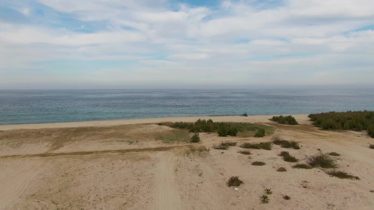 Aerial frame over pale sand and low green shrubs with the Los Barriles beach and blue ocean stretching across the background, with no visible people.