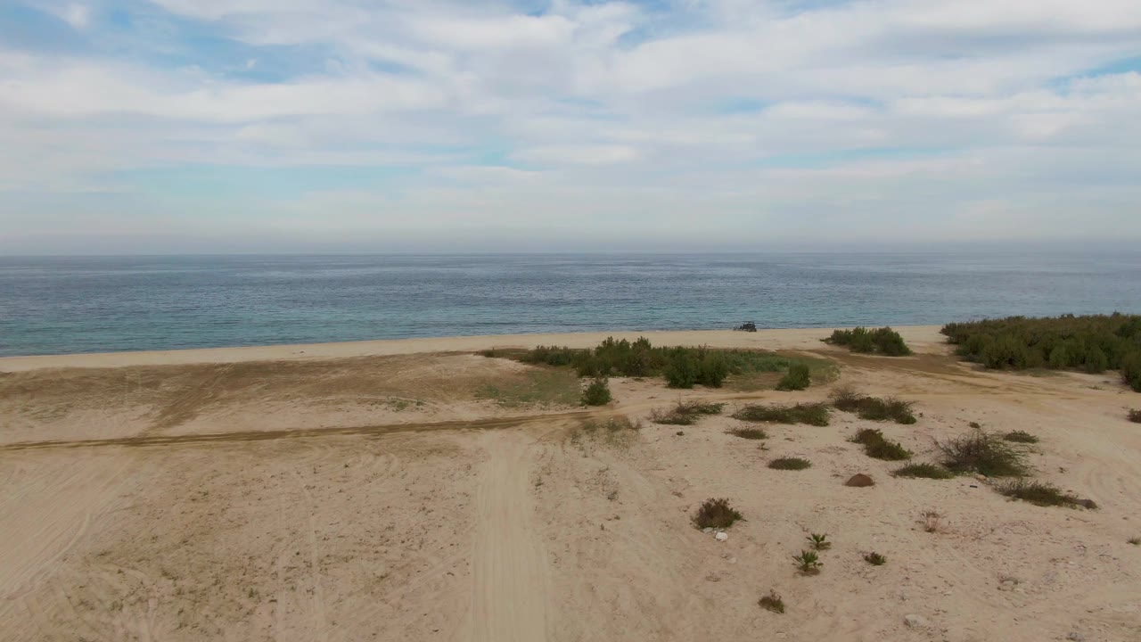 High coastal view in Los Barriles with a pale sandy track leading toward shrub patches, the beach, and a flat blue ocean horizon, with no visible people.