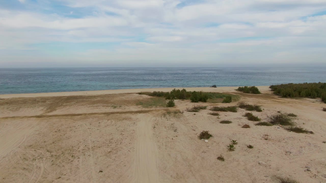 Aerial view across a broad sandy coastal plain in Los Barriles, with scattered shrubs, a narrow beach, blue water, and no visible people.