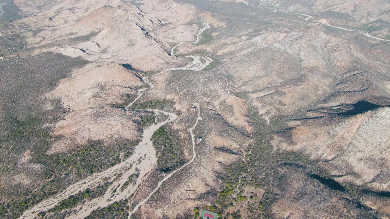 Aerial view over Cabo Pulmo dry hills with scattered green scrub, pale drainage lines, and no visible people at the start of the clip.