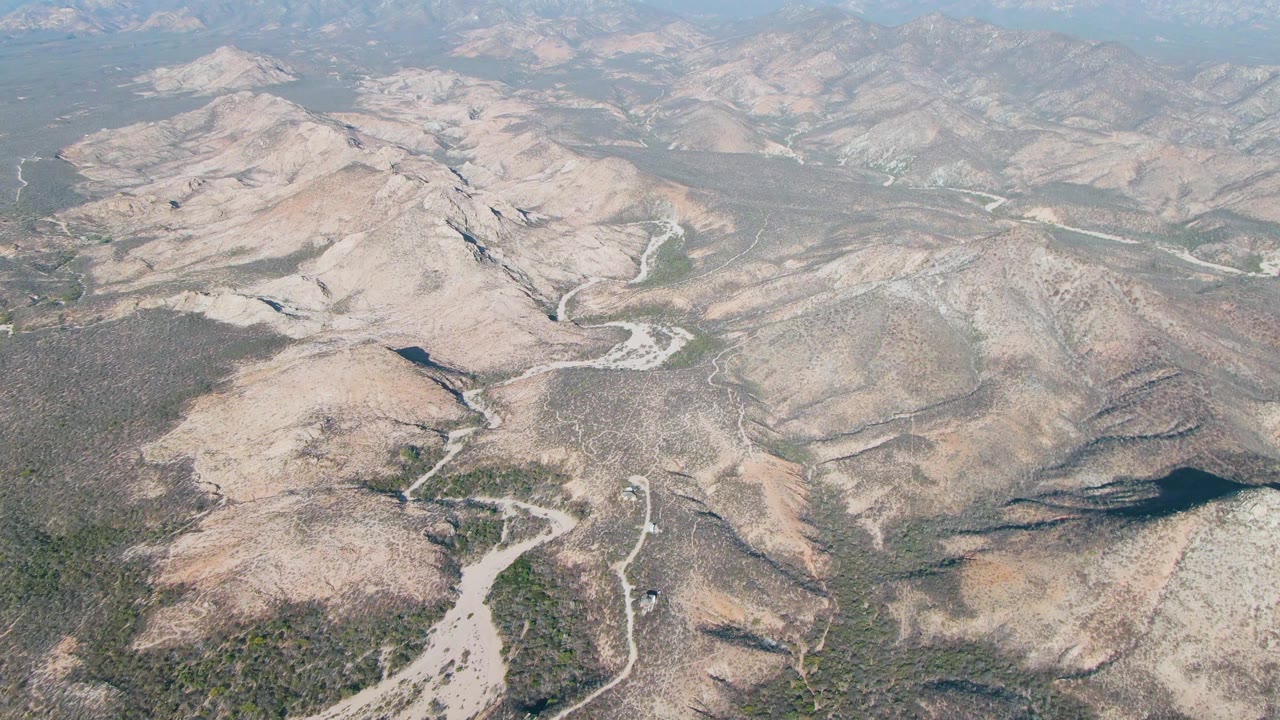 Aerial view in Cabo Pulmo, Baja California Sur, with rounded beige hills, winding pale washes, scattered desert vegetation, a long mountain horizon, and no visible people.