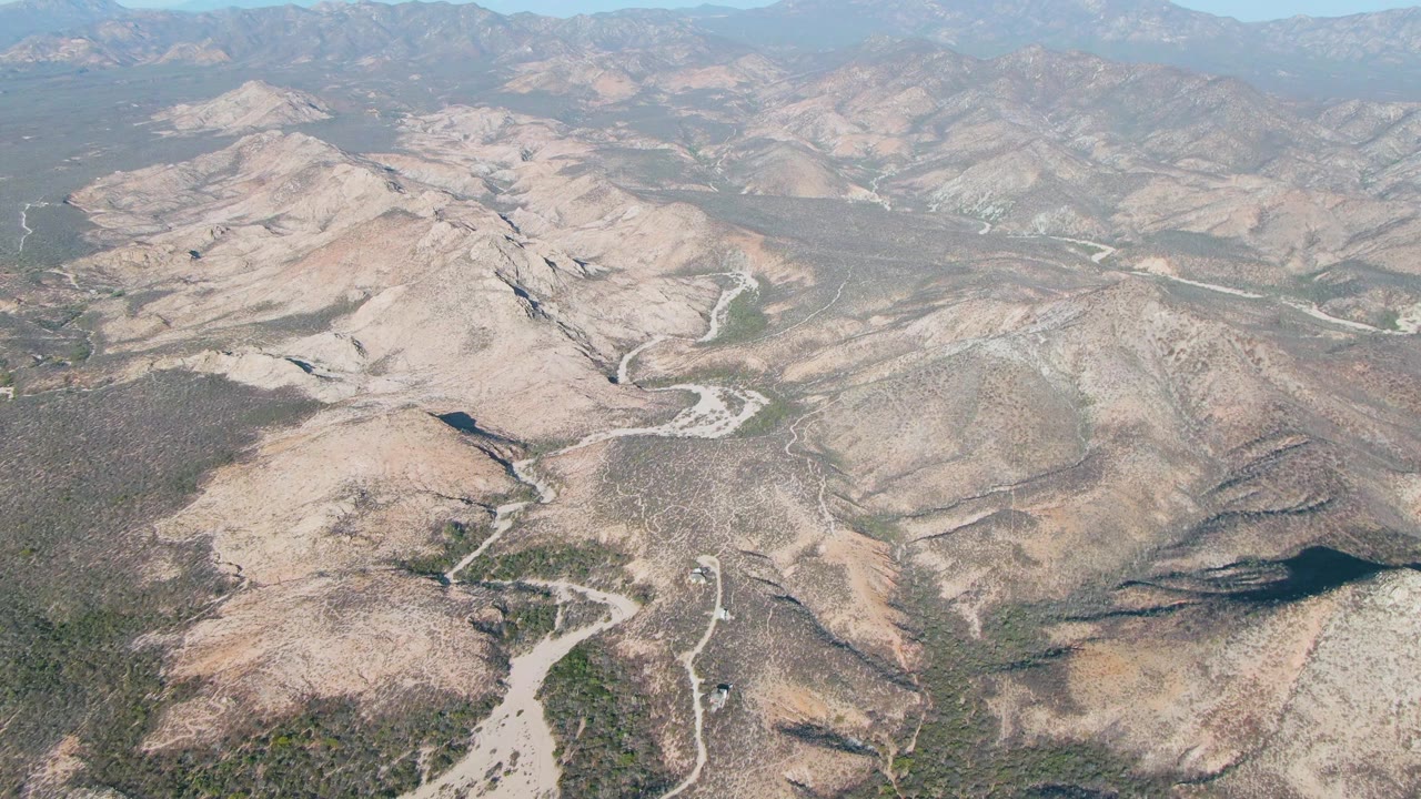 High drone-style view of Cabo Pulmo mountain terrain with tan rocky slopes, pale channels crossing the valley floor, green scrub patches, distant ridgelines, and no visible people.