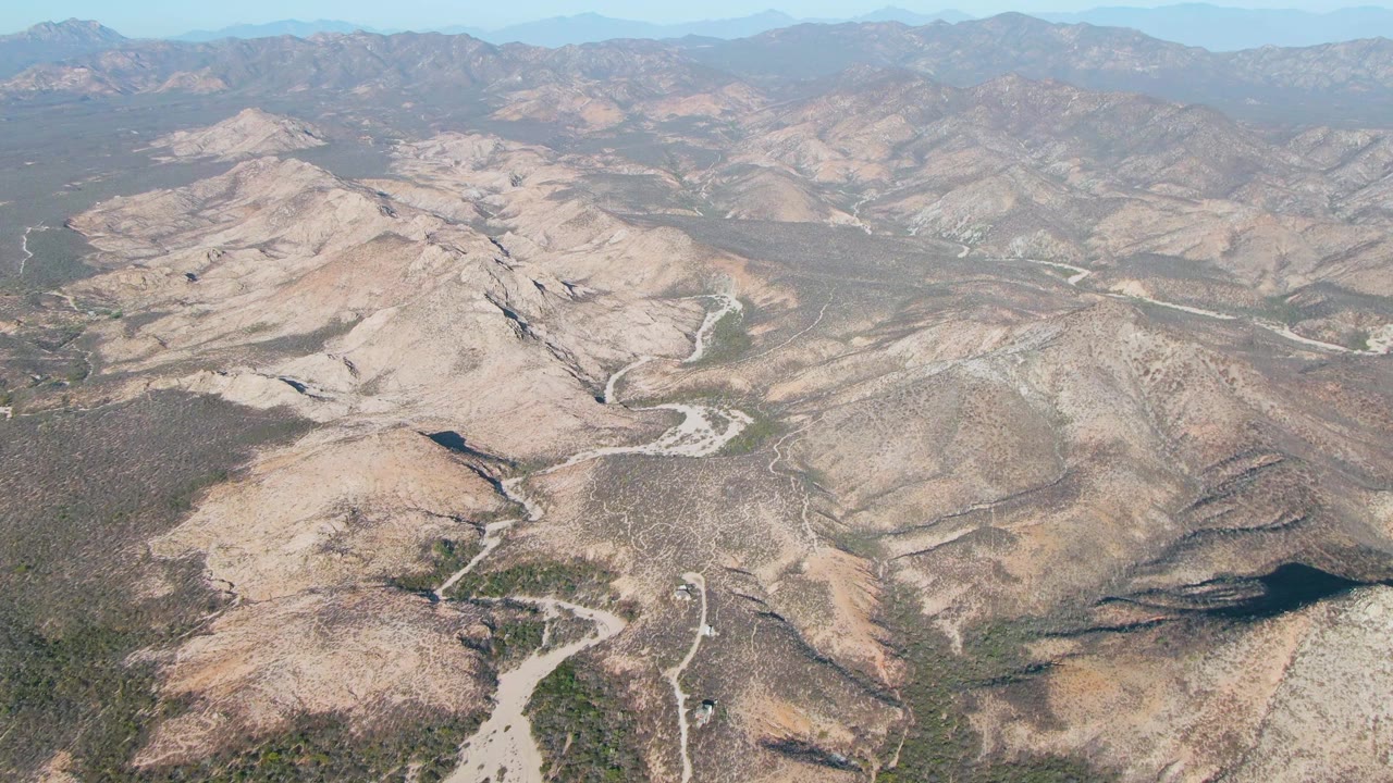 Wide aerial frame over Cabo Pulmo, Baja California Sur, showing layered beige ridges, dry riverbeds, sparse vegetation in low areas, soft haze near the horizon, and no visible people.