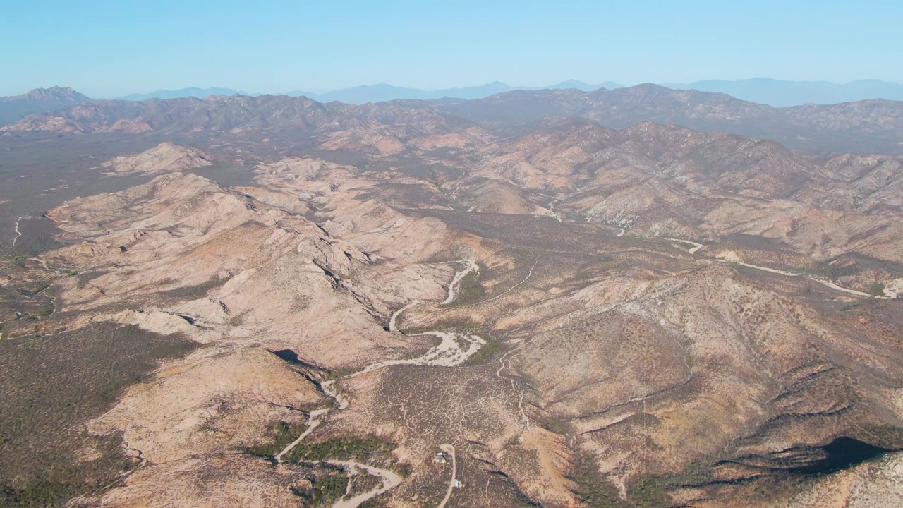 High aerial frame looking across arid mountain folds near Cabo Pulmo, Mexico, where pale roads trace through brown scrub terrain beneath a clear blue sky and no visible people.