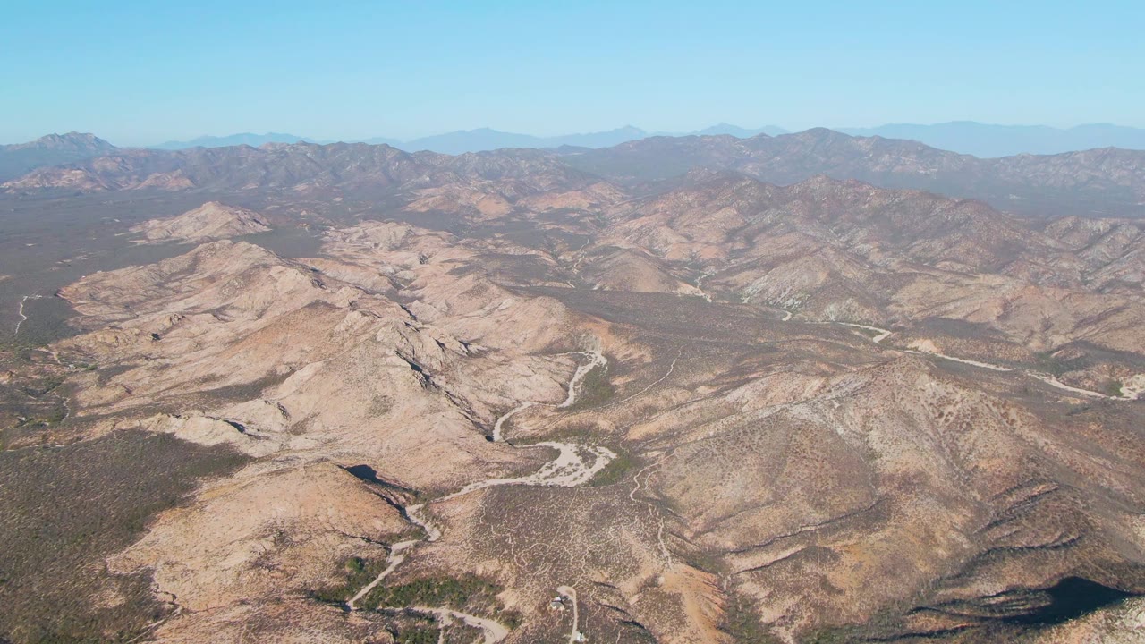 Aerial view of Cabo Pulmo area mountain terrain with a tan ridge in the foreground, winding road lines through the midground, layered hills in the distance, and no visible people.