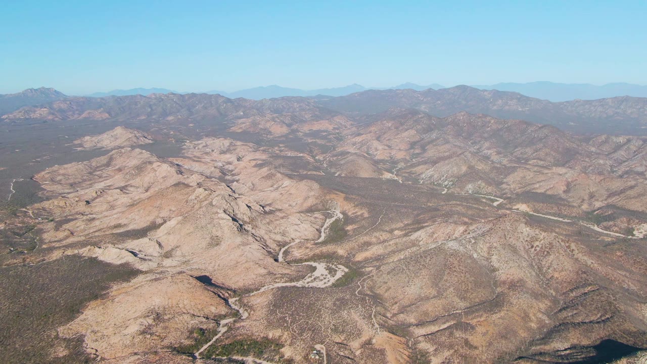 Wide aerial frame over beige hills and branching roads in the Cabo Pulmo East Cape landscape, with dark scrub patches, distant mountain ranges, clear sky, and no visible people.