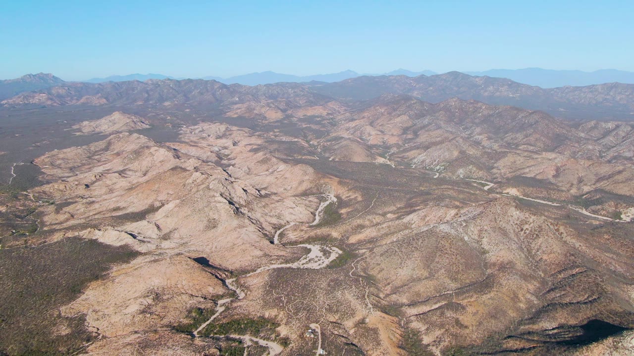 High drone view in Cabo Pulmo with rocky tan ridges, curving dirt tracks, shadowed gullies, distant blue-gray mountains, and no visible people.