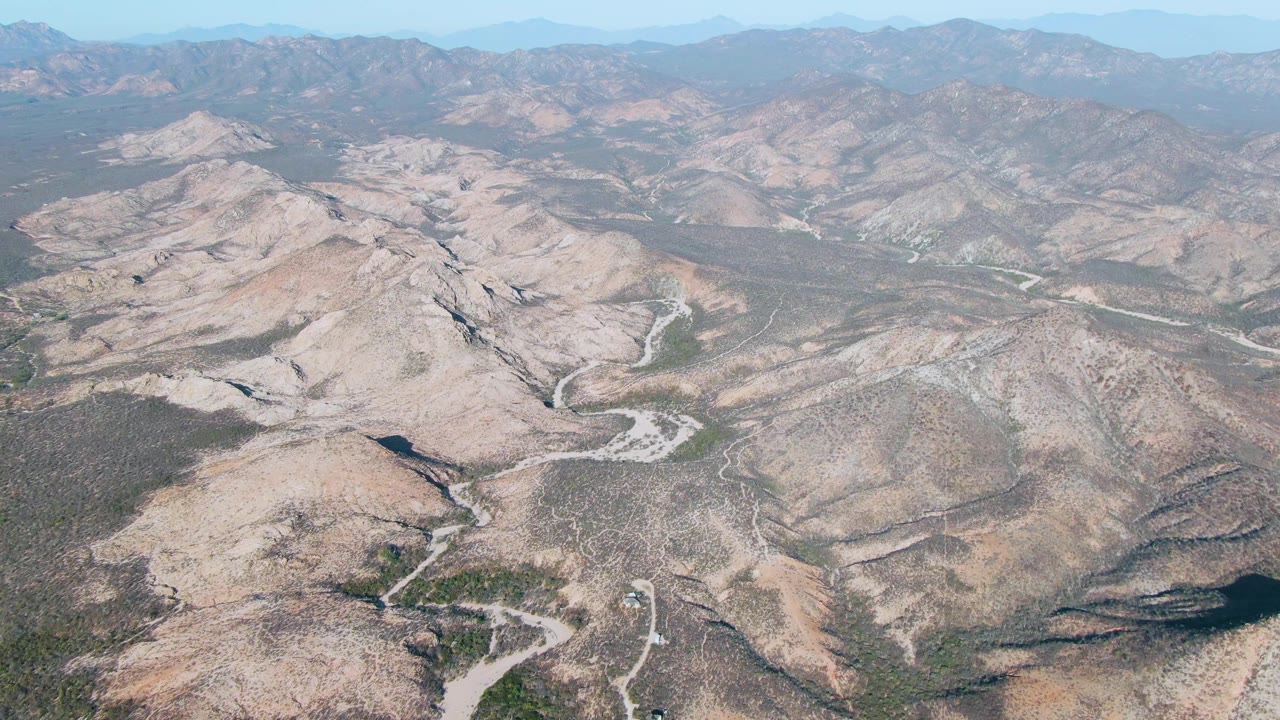 Wide aerial frame over dry hills around Cabo Pulmo, showing winding dirt routes in the midground, layered mountains on the horizon, light haze, and no visible people.