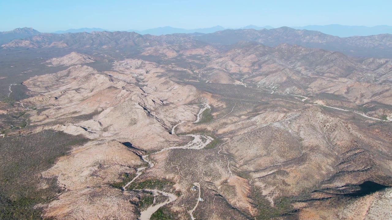 Aerial view of rocky desert hills in Cabo Pulmo, Baja California Sur, with a more open blue sky, dry channels, scattered scrub, and no visible people.