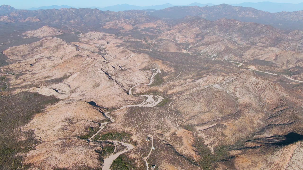 High aerial view over Cabo Pulmo arid mountain terrain, with pale roads cutting through tan hills, distant ridgelines, scattered green vegetation, and no visible people.