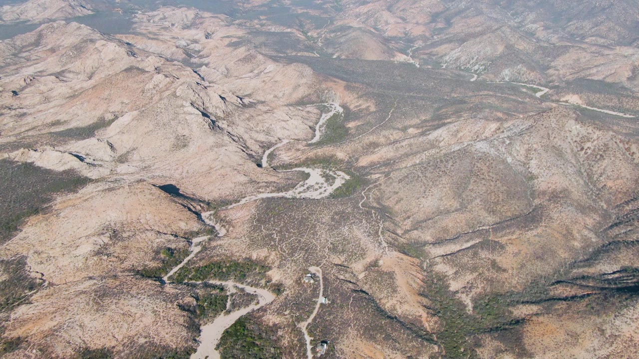 High oblique drone view in Cabo Pulmo showing dry ridges, pale winding tracks through shallow valleys, mottled brown ground, sparse green brush, and no visible people.