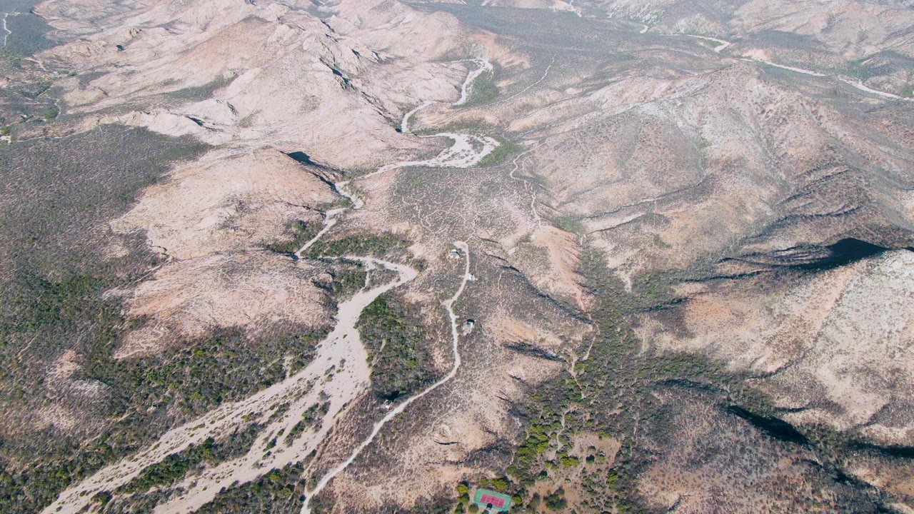 Aerial frame over Cabo Pulmo dry mountain terrain with pale rocky hills, sparse brush, winding dirt roads, a small structure near the lower edge, and no visible people.
