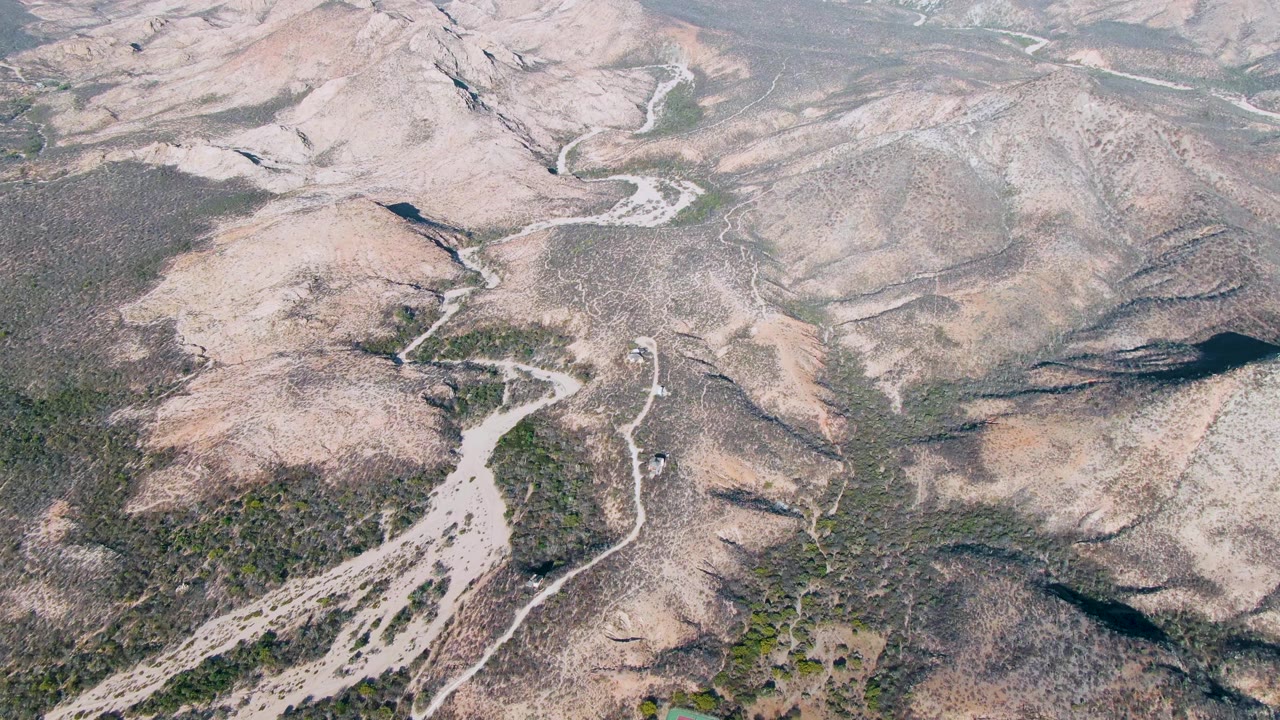 Aerial view over Cabo Pulmo arid hills with winding dirt tracks, dry gullies, patchy green vegetation, rocky tan slopes, and no visible people.