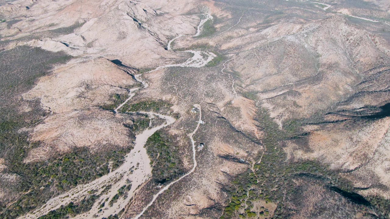 Cabo Pulmo aerial frame showing pale dry hills, branching washes, curving dirt roads, scattered vegetation, and no visible people across the terrain.