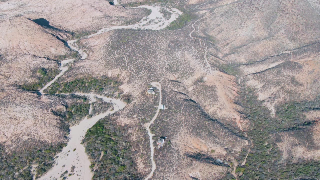 Aerial frame in Cabo Pulmo, Baja California Sur with vehicles continuing along a curved pale road beside darker scrub and dry eroded slopes, with no visible people.