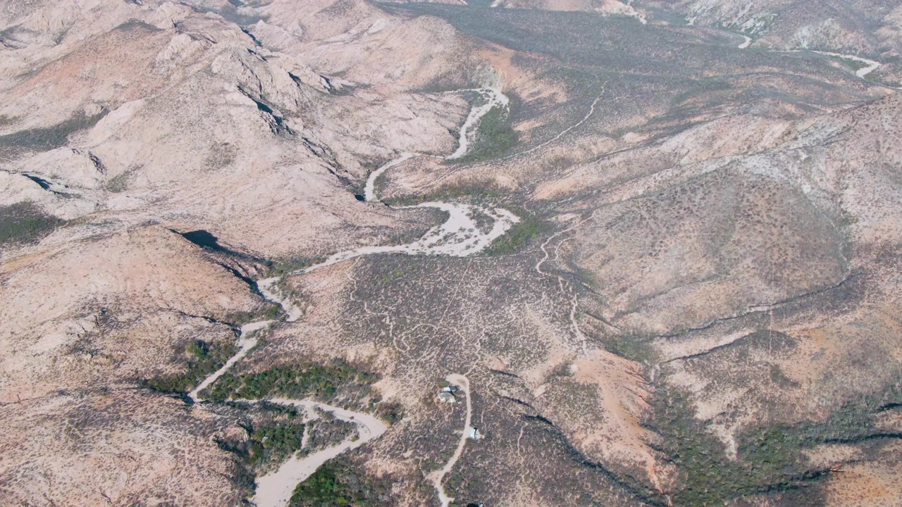 Aerial frame over Cabo Pulmo arid hills in Baja California Sur, with a small vehicle visible on a pale winding road and no visible people.