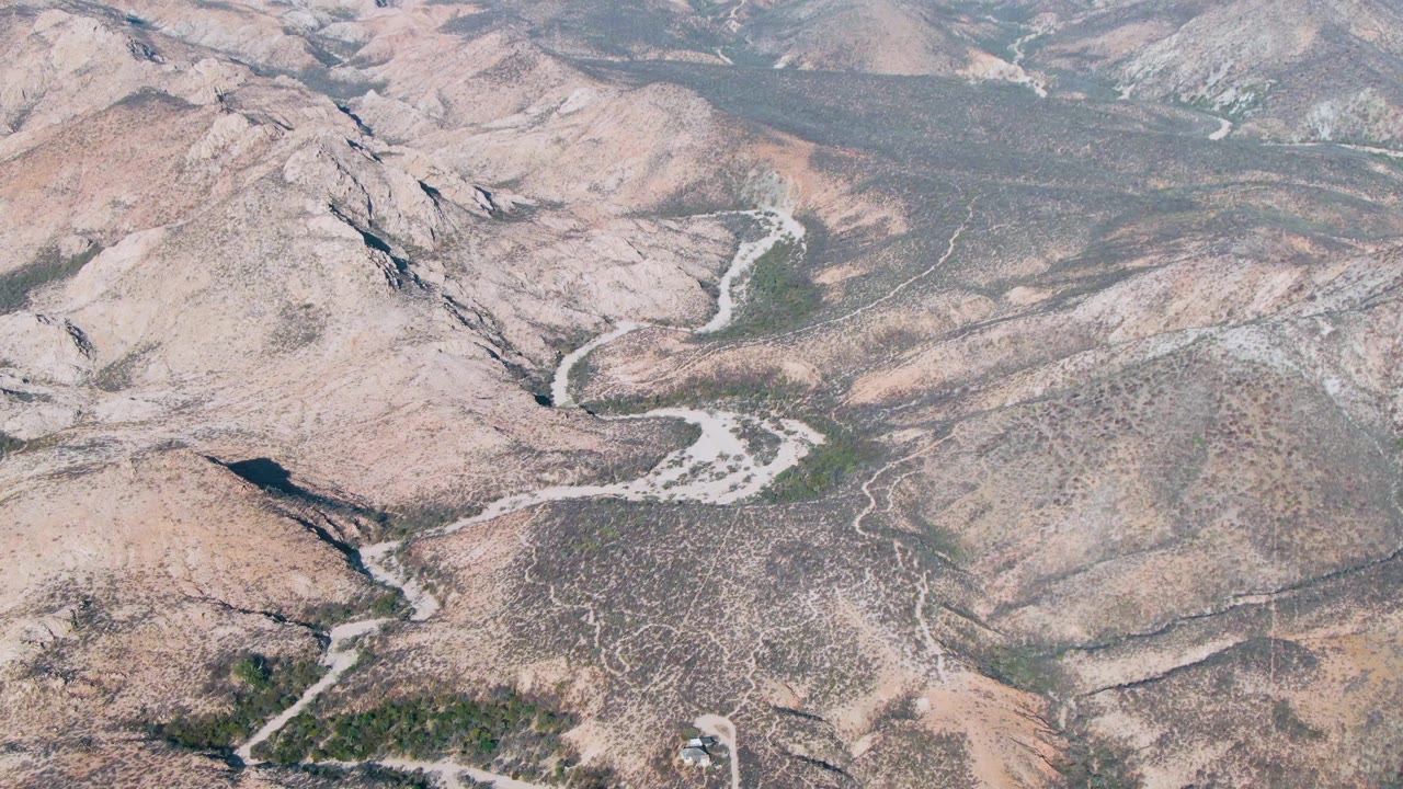 Aerial frame over dry Cabo Pulmo hillside terrain, showing curved dirt tracks, rocky ridges, scattered green patches, and no visible people.