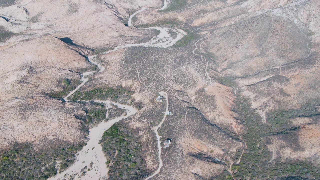 Cabo Pulmo aerial frame with rounded beige hills, branching dry stream channels, darker drainage cuts, scattered scrub, and no visible people.