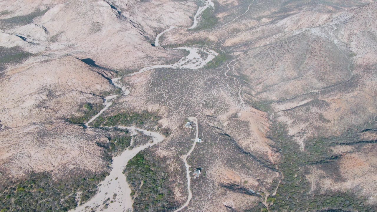 Aerial frame over Cabo Pulmo terrain where a pale dry riverbed curves below rocky hills and sparse green scrub, with no visible people.