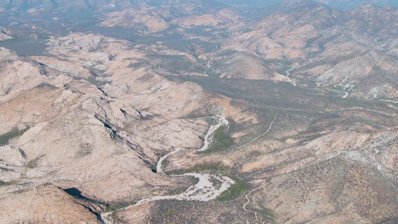 High aerial view near Cabo Pulmo, Baja California Sur, showing beige desert ridges, a pale winding channel, sparse green vegetation, distant mountains, clear sky, and no visible people.