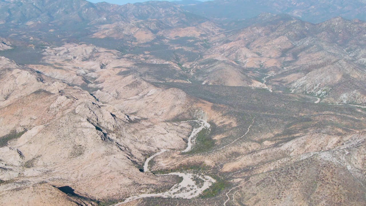 Aerial desert valley with rocky ridges, dry washes, and distant hazy mountains.