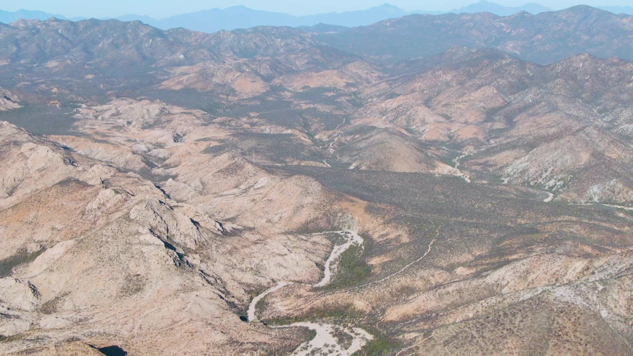 Aerial view near Cabo Pulmo, Baja California Sur, showing a dry mountain basin, pale curving riverbed, layered brown ridgelines, scattered green scrub, clear sky, and no visible people.