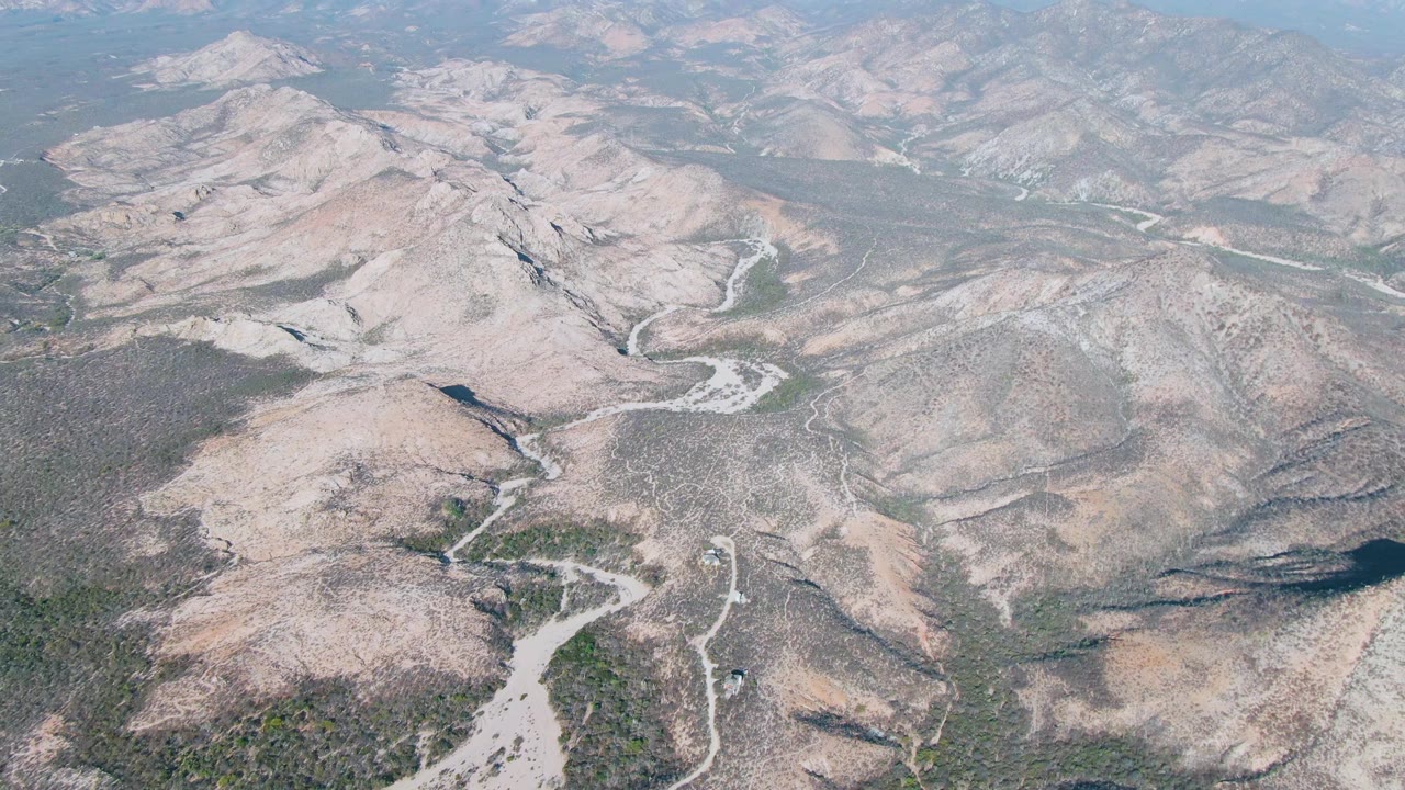Drone-height frame over Cabo Pulmo dry hills, showing the winding wash more centrally between scrub-covered slopes and no visible people.