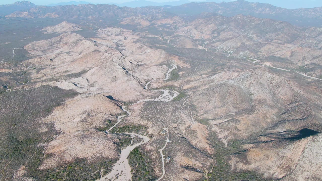 Wide aerial view in Cabo Pulmo with layered arid hills, a winding pale drainage line, distant mountain ridges, and no visible people.