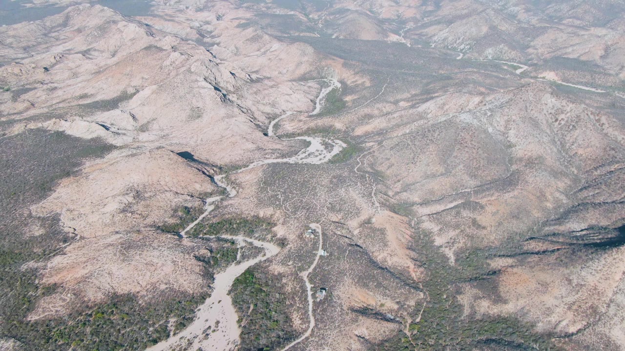 Early aerial frame over Cabo Pulmo area mountains in Baja California Sur, showing pale rocky slopes, dry valleys, winding dirt roads, sparse vegetation, hazy sky, and no visible people.
