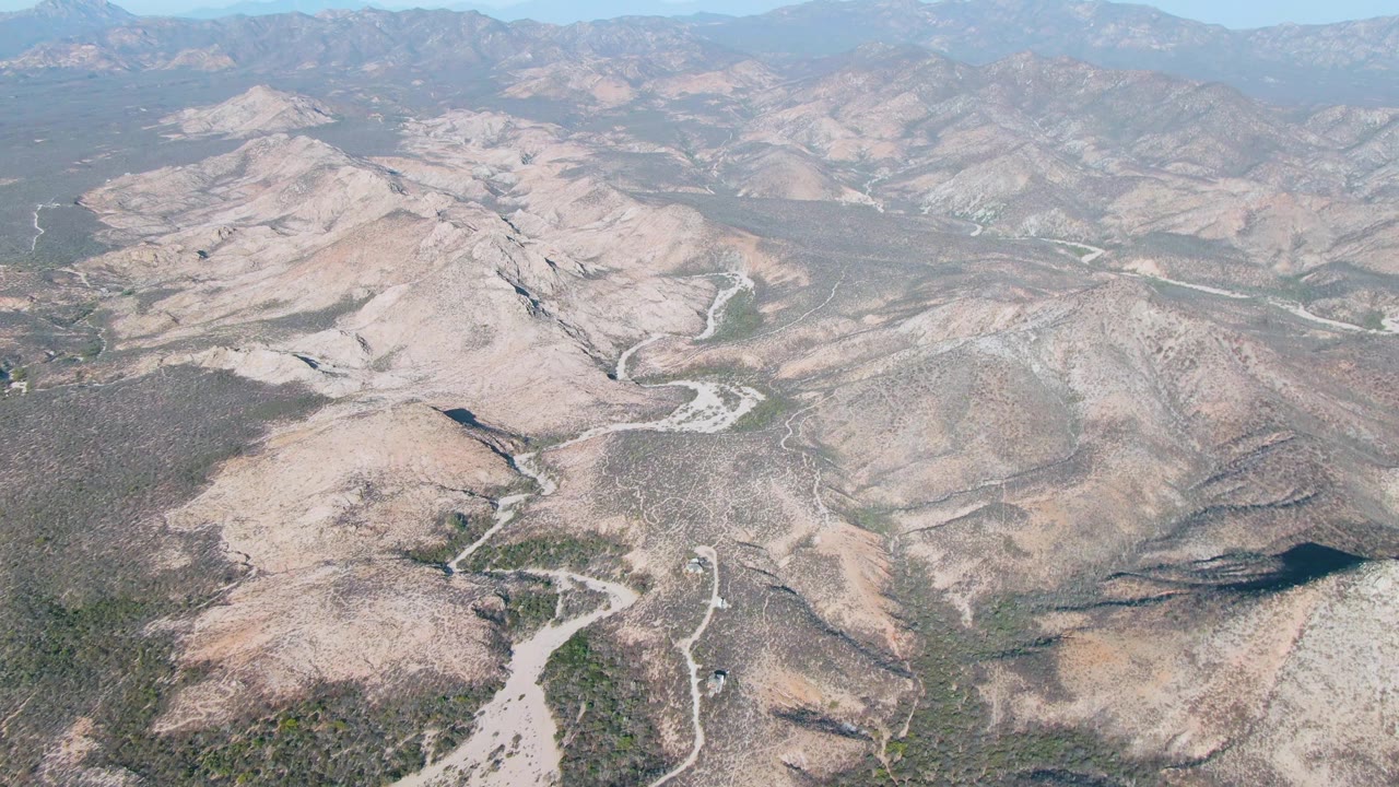 Mid-clip aerial frame over Cabo Pulmo East Cape terrain, with a central rocky hill, branching dry washes, curved dirt roads, distant ridgelines, and no visible people.