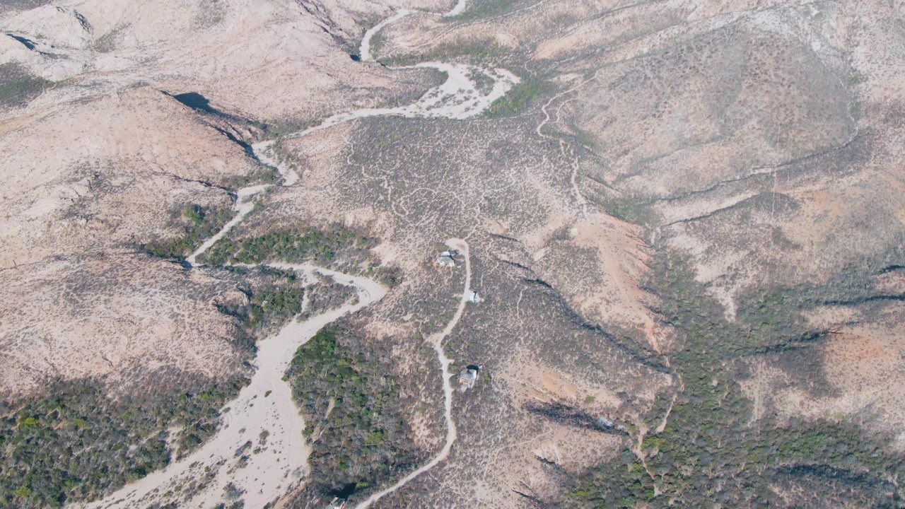 Aerial view of Cabo Pulmo arid terrain with the road bending through pale hills and a small vehicle visible near the route, with no visible people.
